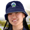 A white bucket hat with a navy sandwich trim and a logo, worn by a smiling person outdoors.