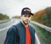 A navy twill cap with a logo, worn by a man in a grey jacket, standing on a road with autumn foliage in the background.