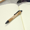 A black plastic pen with a cork grip rests on an open notebook with lined pages. It features a logo.