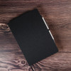 A black A5 notebook with a textured cover and an attached pen on a wooden surface. It features a logo on the front.