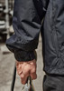 A close-up of a person's arm wearing a black packable rain jacket, holding a rope. The jacket features an adjustable cuff.