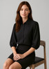 A black 3/4 sleeve blouse with a collared neckline, styled with a fitted skirt, displayed on a seated model.