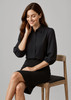 A black 3/4 sleeve blouse worn by a woman sitting on a wooden chair, paired with a black skirt.