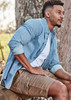A man sits against a tree wearing a light blue button-up shirt and khaki work shorts, showcasing a casual style.