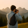 A person wearing a denim jacket holds a camera, showcasing a dark blue sling bag against a mountain landscape.