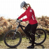 A woman in a red lightweight jacket and helmet is riding a black mountain bike in a grassy area.