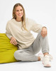 A unisex cream crew neck sweater paired with grey sweatpants, worn by a smiling model seated on a green cushion.