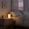 A foldable lantern in white emits a warm glow on a bedside table next to a glass of water and a small plant.