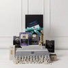 A gourmet gift pack featuring assorted food items, a stylish grey throw, and a bottle, all presented in a black box.