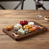 A slate cheeseboard with a wooden frame, featuring assorted cheeses, nuts, grapes, and garnishes. A glass of wine is nearby.