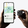 A hand holds a small black Bluetooth tracker next to a smartphone displaying a map and tracking interface.