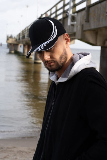 A man wearing a black cap with a white checkered design and a black jacket stands near water.