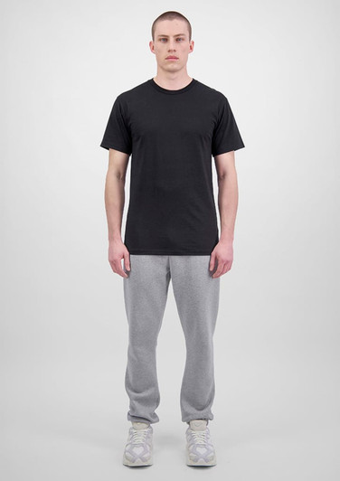 A person wears a black t-shirt and grey track pants, standing against a plain background. The outfit features branding.
