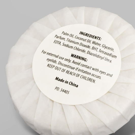 A white circular soap wrapped in paper, labelled with ingredients and warnings, features a logo.