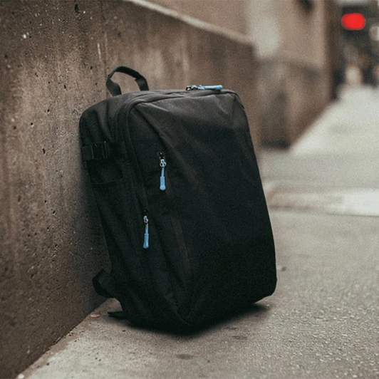 A black travel backpack with blue zipper pulls leaning against a concrete wall. It features multiple compartments.