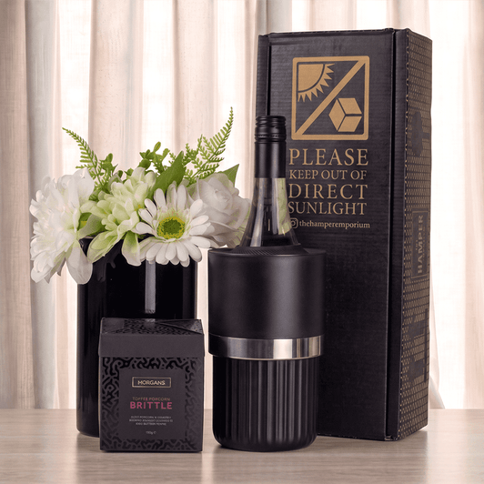 A black wine bottle cooler with silver band, a box of brittle, and a vase with white flowers, arranged on a table.