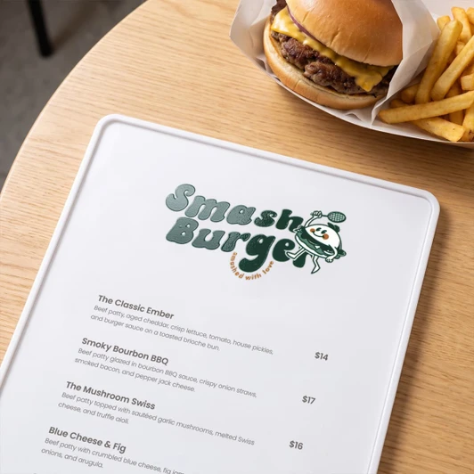 A menu on a wooden table featuring various burger options alongside a cheeseburger and fries in the background.
