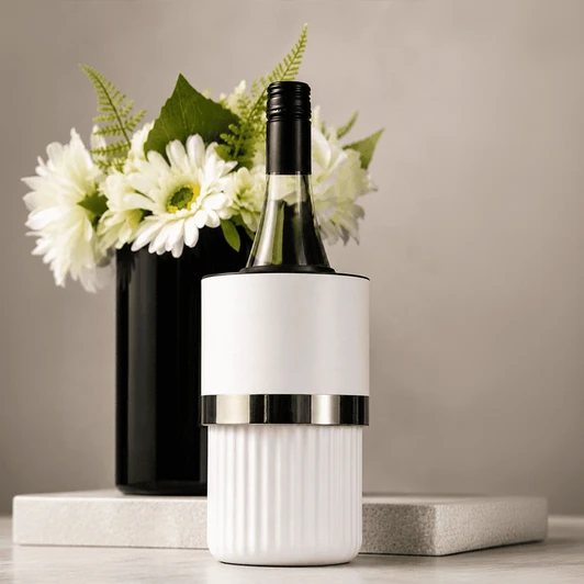 A white wine bottle cooler with a silver band, placed on a marble surface beside a black vase with white flowers.