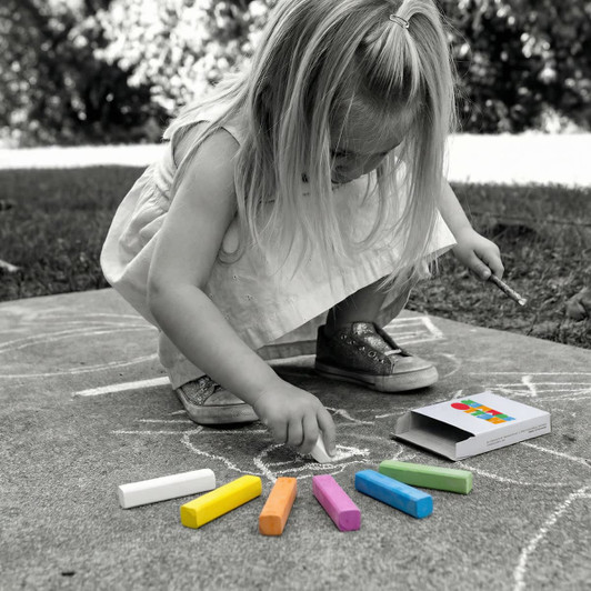 A child uses colourful chalk sticks, including white, orange, pink, green, and purple, from an open box on the ground.