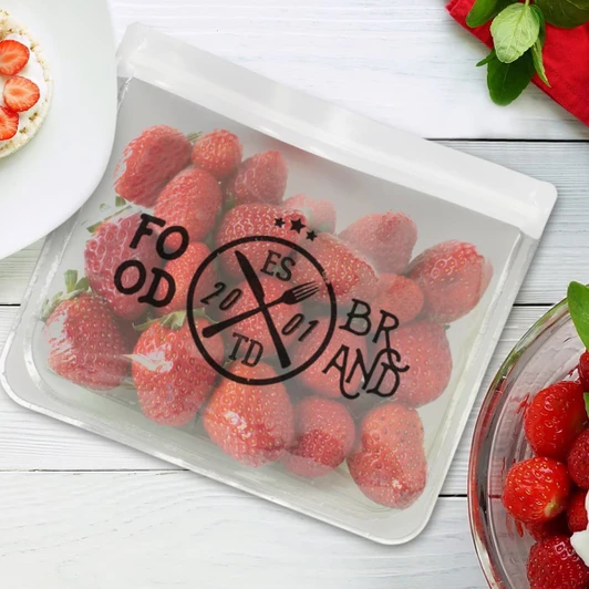 A transparent reusable pouch filled with fresh strawberries, featuring a circular logo design.