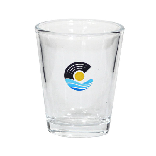 A clear glass shot glass featuring a logo with blue waves and a sun design.