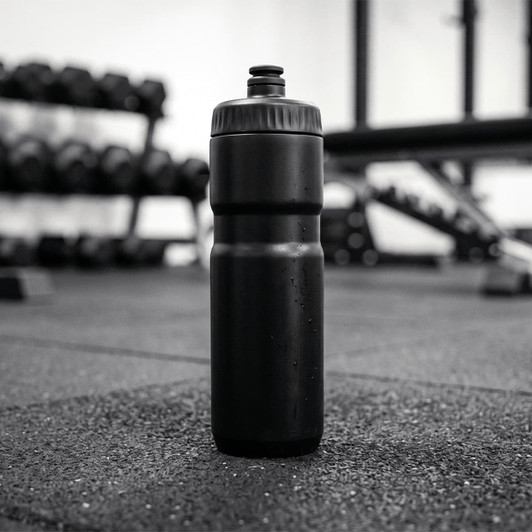 A drink bottle in black, standing upright on a gym floor with weights in the background.