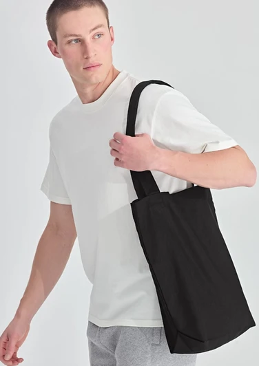 A man in a white T-shirt carries a black canvas tote bag with a shoulder strap. The bag has a simple design.