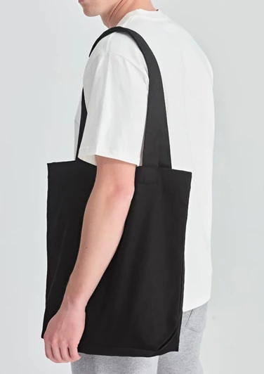 A black canvas tote bag is worn over the shoulder by a person in a white t-shirt and grey sweatpants.