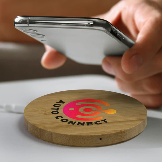 A round bamboo wireless charger in a natural finish, with a logo on the surface, beside a smartphone.