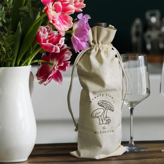 A cream cotton drawstring bag with a logo, holding a wine bottle, next to a vase of pink flowers and glasses.