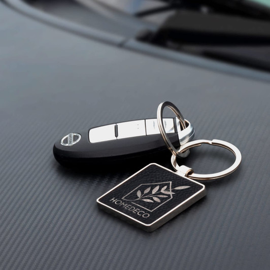 A square silver key ring with a logo, attached to a car key, placed on a black textured surface.