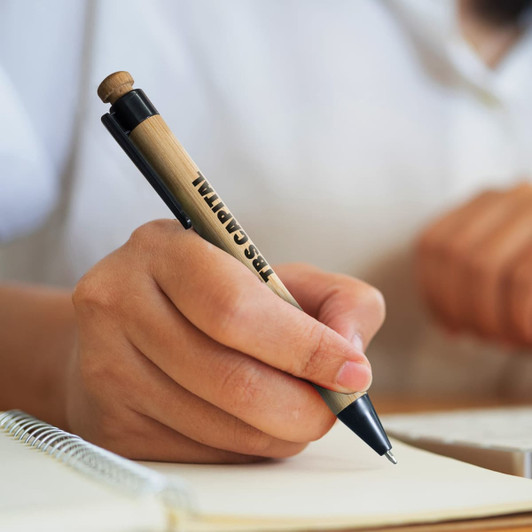 A bamboo pen with a black tip, held in a hand, writing on a notebook. It features a branding logo.