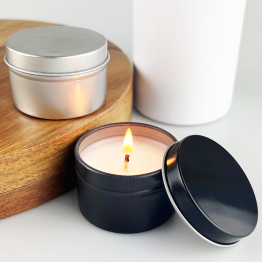 A black tin candle with a lit wick beside a silver tin on a wooden surface.