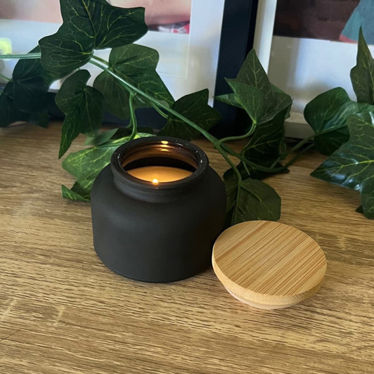 A black candle jar with a bamboo lid, placed on a wooden surface, surrounded by green ivy.