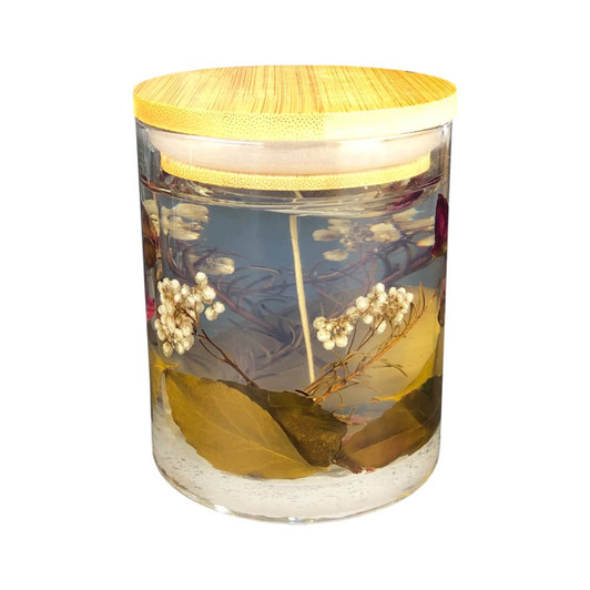 A floral candle in a clear glass jar, featuring dried flowers and leaves, topped with a bamboo lid.