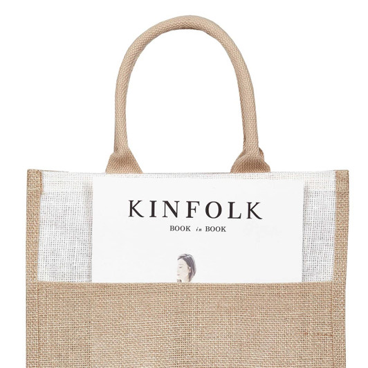 A jute tote bag with sturdy handles featuring a book partially visible in the front pocket. The bag is a natural tan colour.