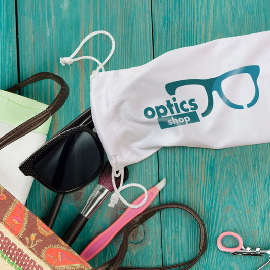 A white microfibre pouch with a logo, surrounded by sunglasses, scissors, and cosmetic items on a wooden surface.
