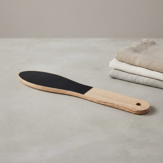 A wooden foot file with a light timber handle and a black abrasive surface, placed beside neatly folded cloths.