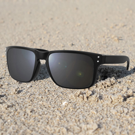 A pair of black sunglasses resting on sandy ground, featuring a simple design and grey lenses.