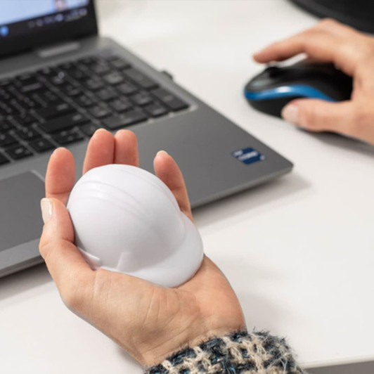 A hand holds a white stress hard hat, resting on a desk with a laptop and computer mouse nearby.
