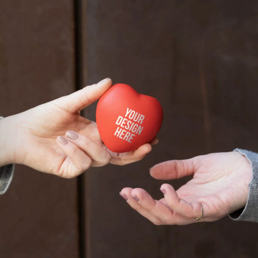 A red heart-shaped stress ball is being handed from one hand to another. The product features a blank area for custom design.