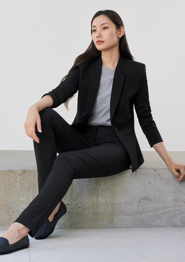 A woman sits casually in a black blazer and matching black pants, paired with a grey top and black shoes.
