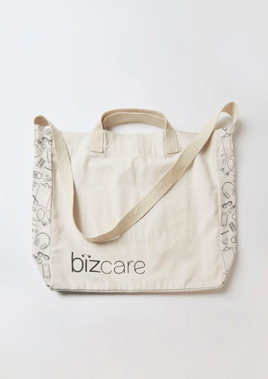 A cream tote bag with a strap, featuring a logo and patterned detailing along the edges.