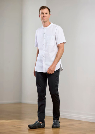 A model wearing a white short-sleeve chef shirt and black chef pants, standing in a light-filled room.
