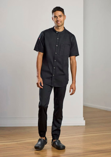 A model wearing black jogger pants and a matching short-sleeve chef shirt, standing in an empty room.