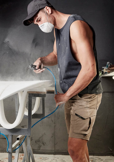 A sleeveless black tee worn by a man working on a surfboard in a workshop, wearing a mask and cap.