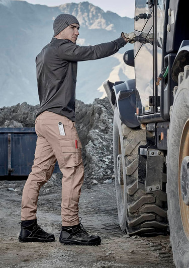 A man wearing beige curved cargo pants and a long-sleeve shirt stands next to a truck in a rugged outdoor setting.