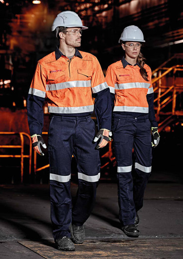 Two individuals wearing high-visibility orange shirts and navy cargo pants walk in an industrial setting, both wearing helmets and gloves.