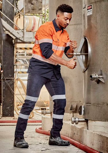 A man in an orange and navy work shirt and blue stretch jeans adjusts equipment, featuring reflective stripes and safety boots.