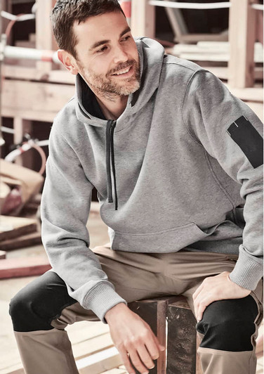 A grey unisex multi-pocket hoodie is worn by a man seated on a wooden bench, with worksite equipment in the background.